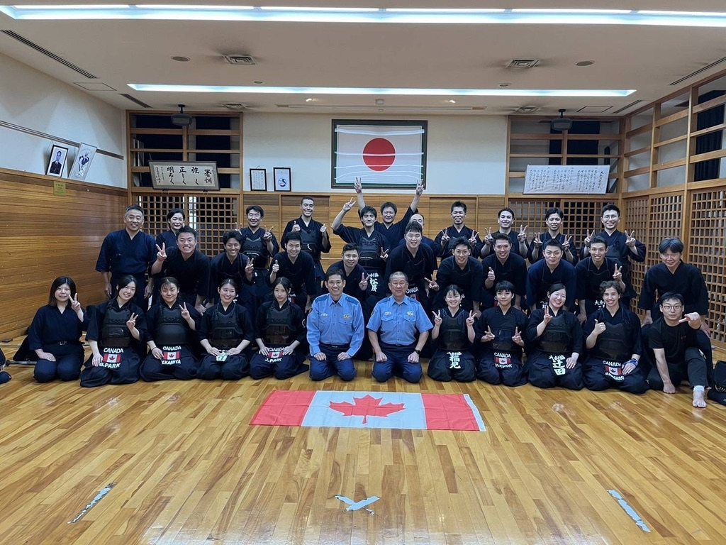 Team Canada and the Osaka Prefectural Police kendo squad posed together in a police dojo, two officers in uniform at the centre and a Canadian flag laid on the floor beneath a Japanese flag