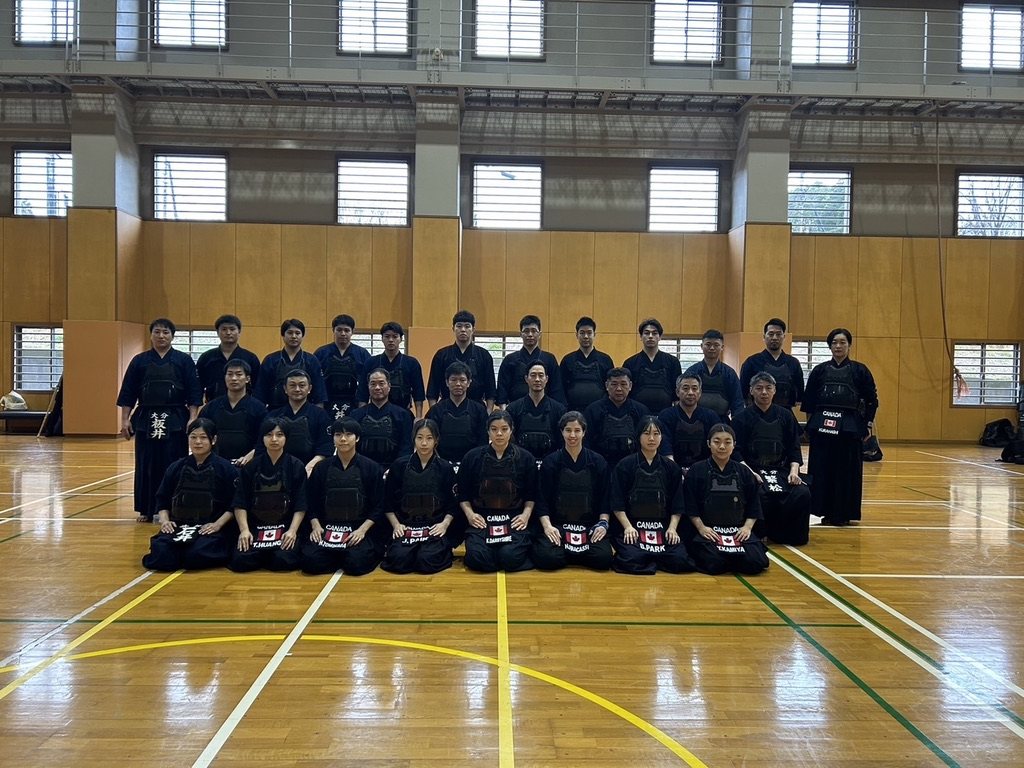 Team Canada posed in rows in bogu alongside the Oita Prefectural Police kendo squad in a large gymnasium