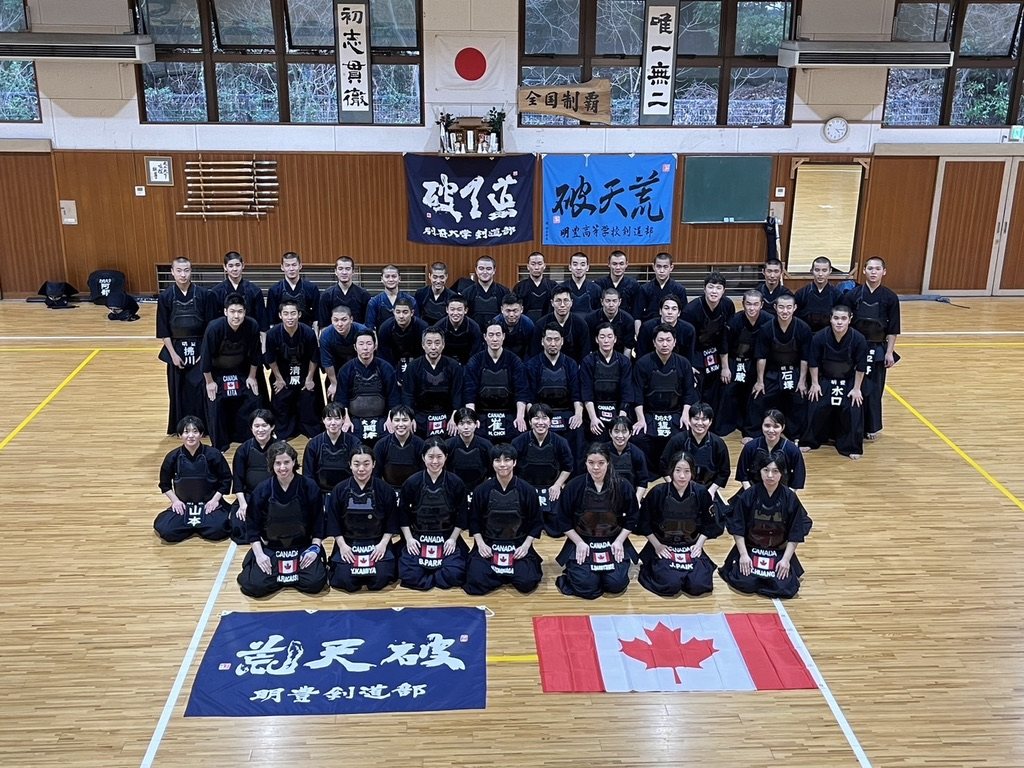 Team Canada and a Japanese high school kendo club posed together in a school dojo, club banners and a Japanese flag on the wall and a Canadian flag laid on the floor