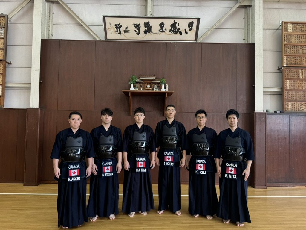 The six-man Team Canada kendo squad standing in a line in a traditional dojo beneath a calligraphy plaque, name plates reading CANADA
