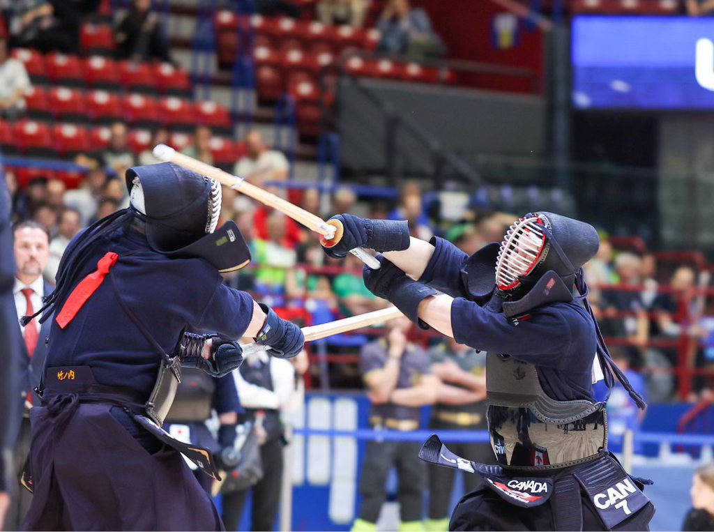 Two kendoka mid-exchange on the competition floor, shinai crossed, a Team Canada player on the right