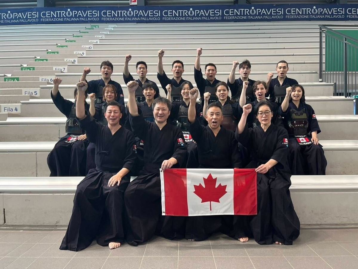 Team Canada kendo squad seated on arena bleachers holding a Canadian flag, fists raised